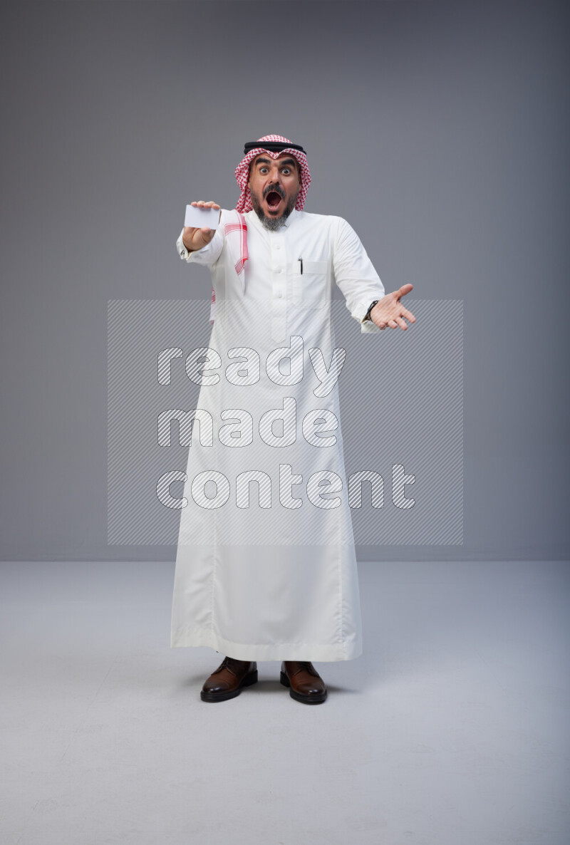 Saudi man Wearing Thob and red Shomag standing holding ATM card on Gray background