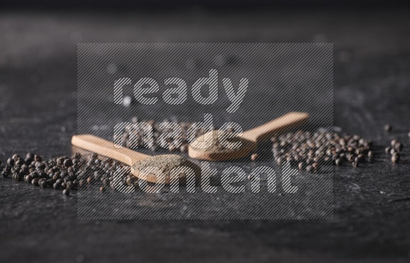 2 wooden spoons full of black pepper powder and black pepper beads spread on a textured black flooring
