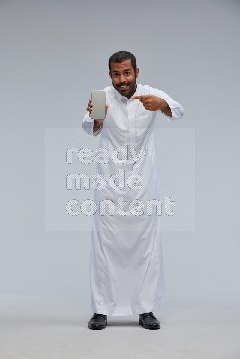 Saudi man wearing Thob standing showing phone to camera on Gray background