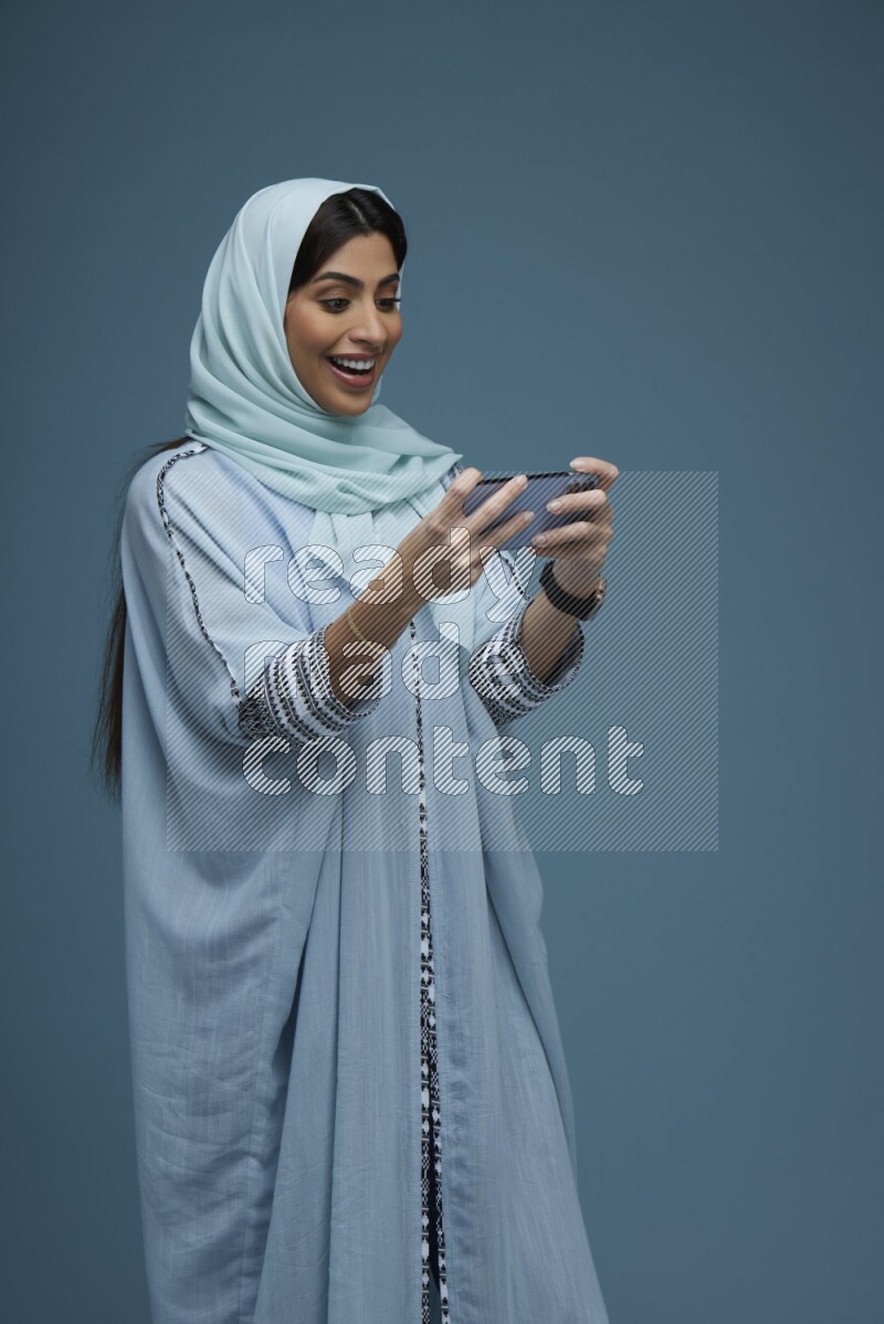 A Saudi woman Playing a Game on her phone on a blue background wearing a blue Abaya with hijab