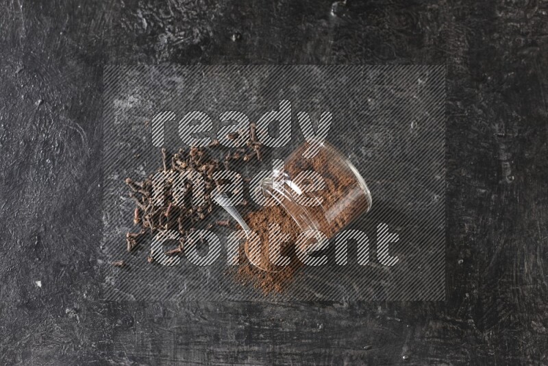 A flipped glass jar and metal spoon full of cloves powder with cloves spread on a textured black flooring