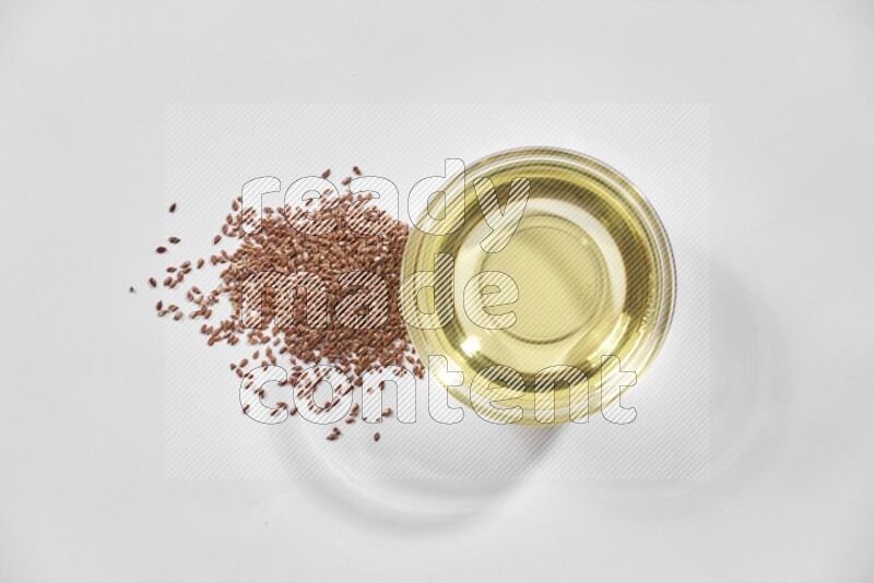 A glass bowl full of flaxseeds oil and flaxseeds beside it on a white flooring