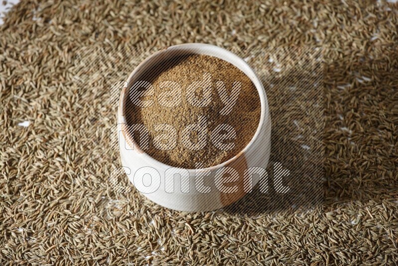 وعاء بيج ممتلئ ببودرة الكمون مع حبوب الكمون تحت الوعاء على خلفية بيضاء