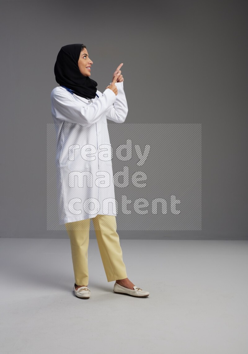 Saudi woman wearing lab coat with stethoscope standing interacting with the camera on Gray background