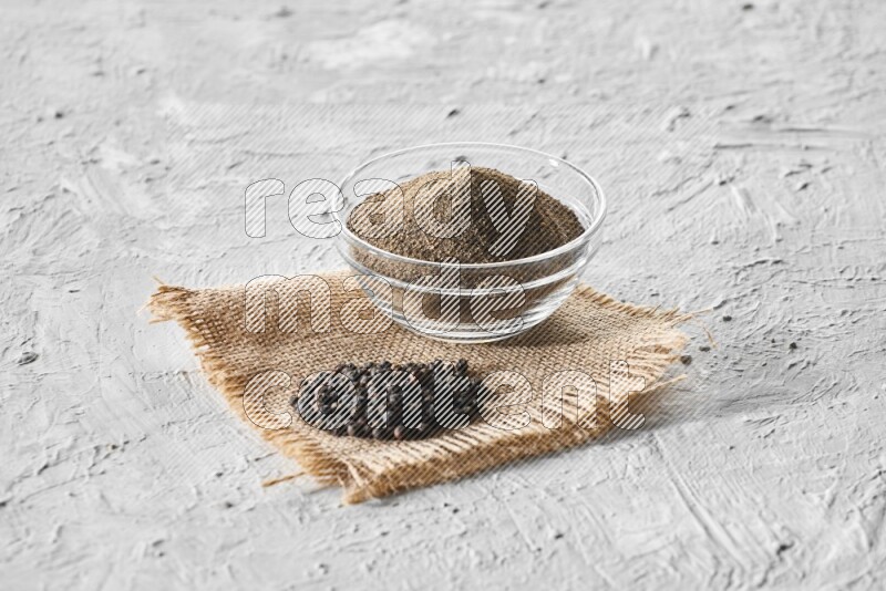 A glass bowl full of black pepper powder and black pepper beads on burlap fabric on textured white flooring