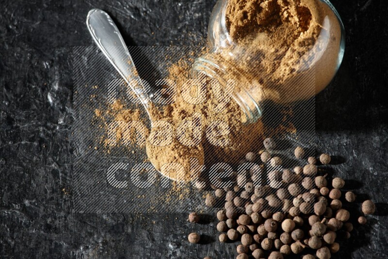 A flipped glass spice jar and metal spoon full of allspice powder and powder spilled out of it with whole balls on a textured black flooring