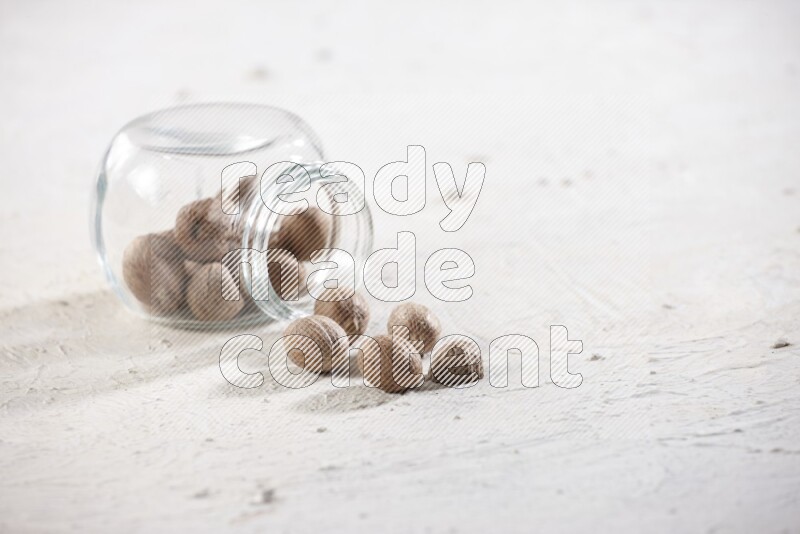 A glass spice jar full of whole nutmeg seeds flipped and the seeds spread out on a textured white flooring