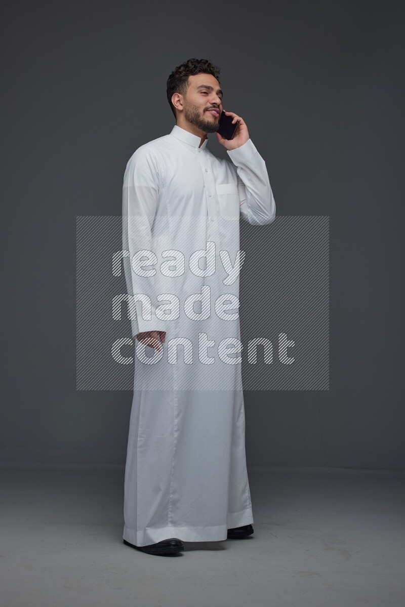 A Saudi man wearing Thobe and talking in the phone while standing and making different poses eye level on a gray background