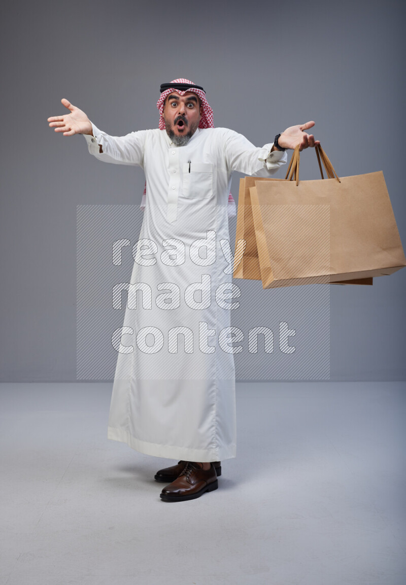Saudi man Wearing Thob and red Shomag standing holding shopping bag on Gray background