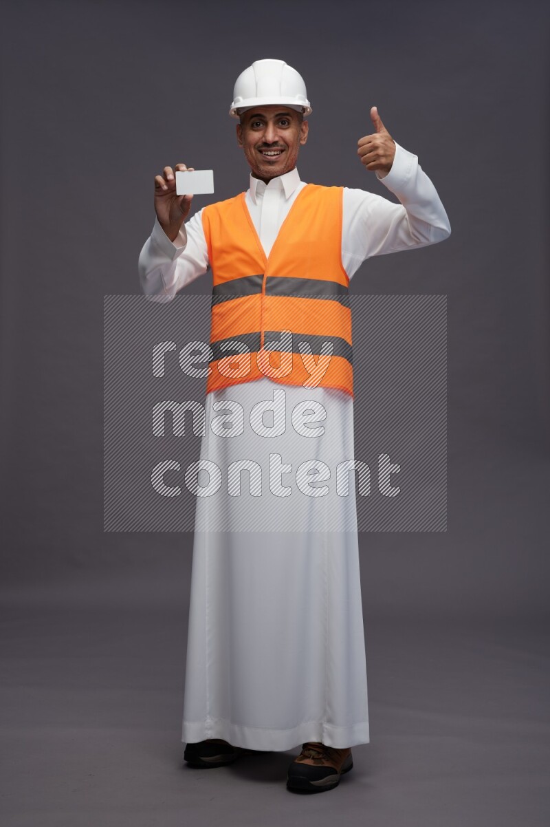 Saudi man wearing thob with engineer vest standing holding ATM card on gray background