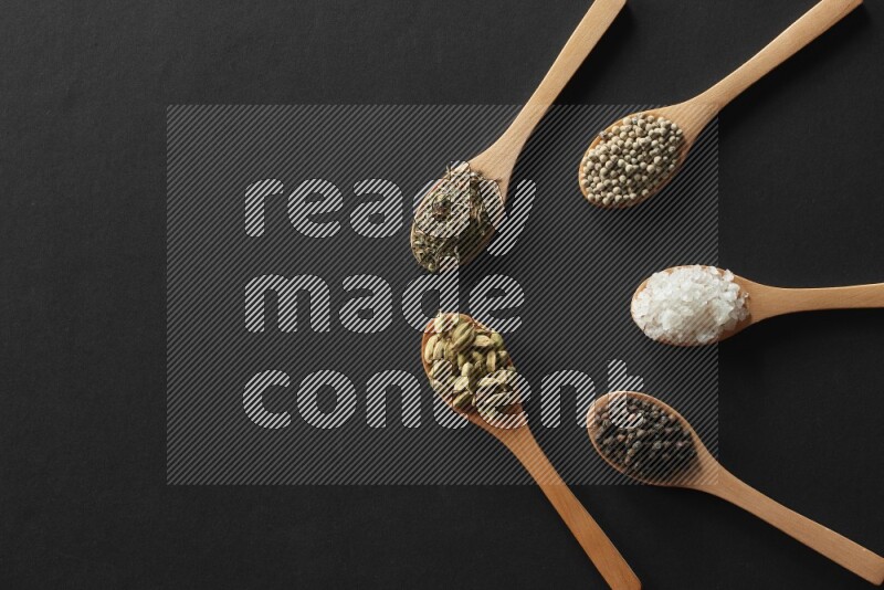 wooden spoons filled with white peppers, cloves, cardamom, salt, black peppers and basil on black flooring