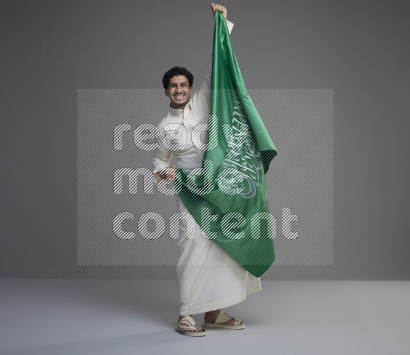 A Saudi man standing wearing thob holding big Saudi flag on gray background