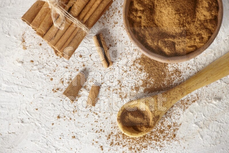Cinnamon sticks stacked and bounded beside a wooden bowl full of cinnamon powder and a wooden spoon full of powder on white background