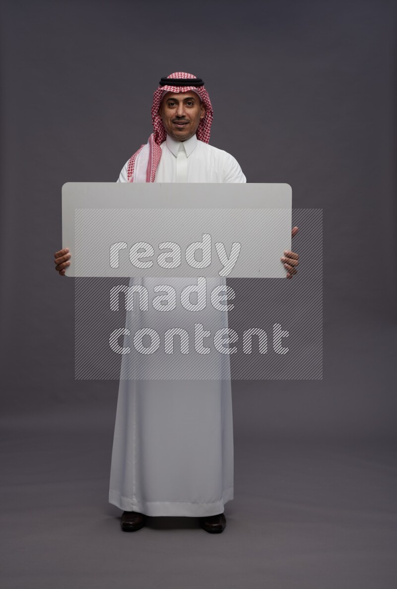 Saudi man wearing thob and shomag standing holding board on gray background