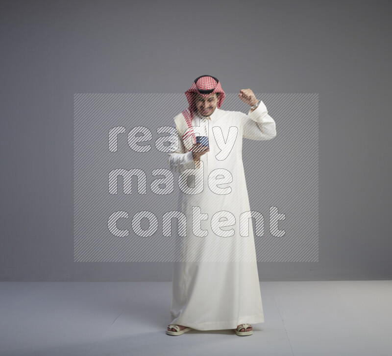 A  Saudi man standing wearing thob and red shomag texting on phone on gray background