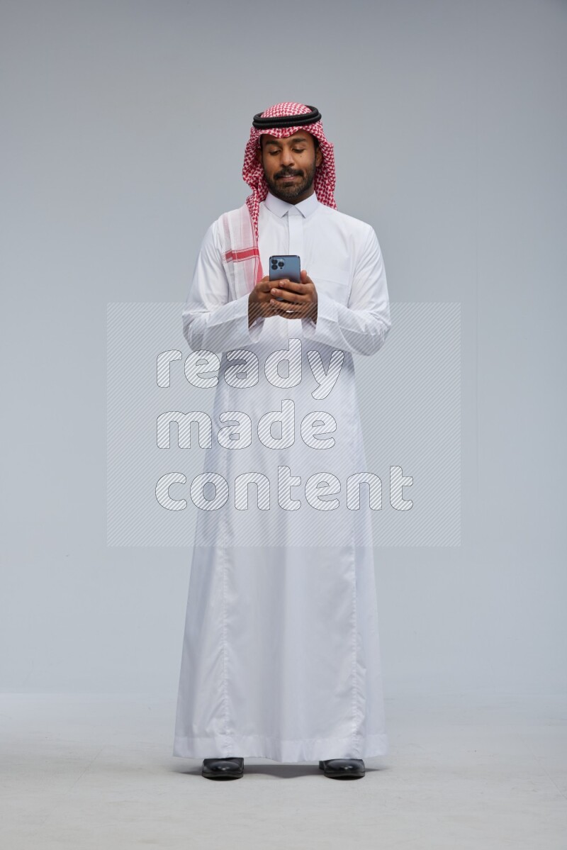Saudi man Wearing Thob and shomag standing texting on phone on Gray background