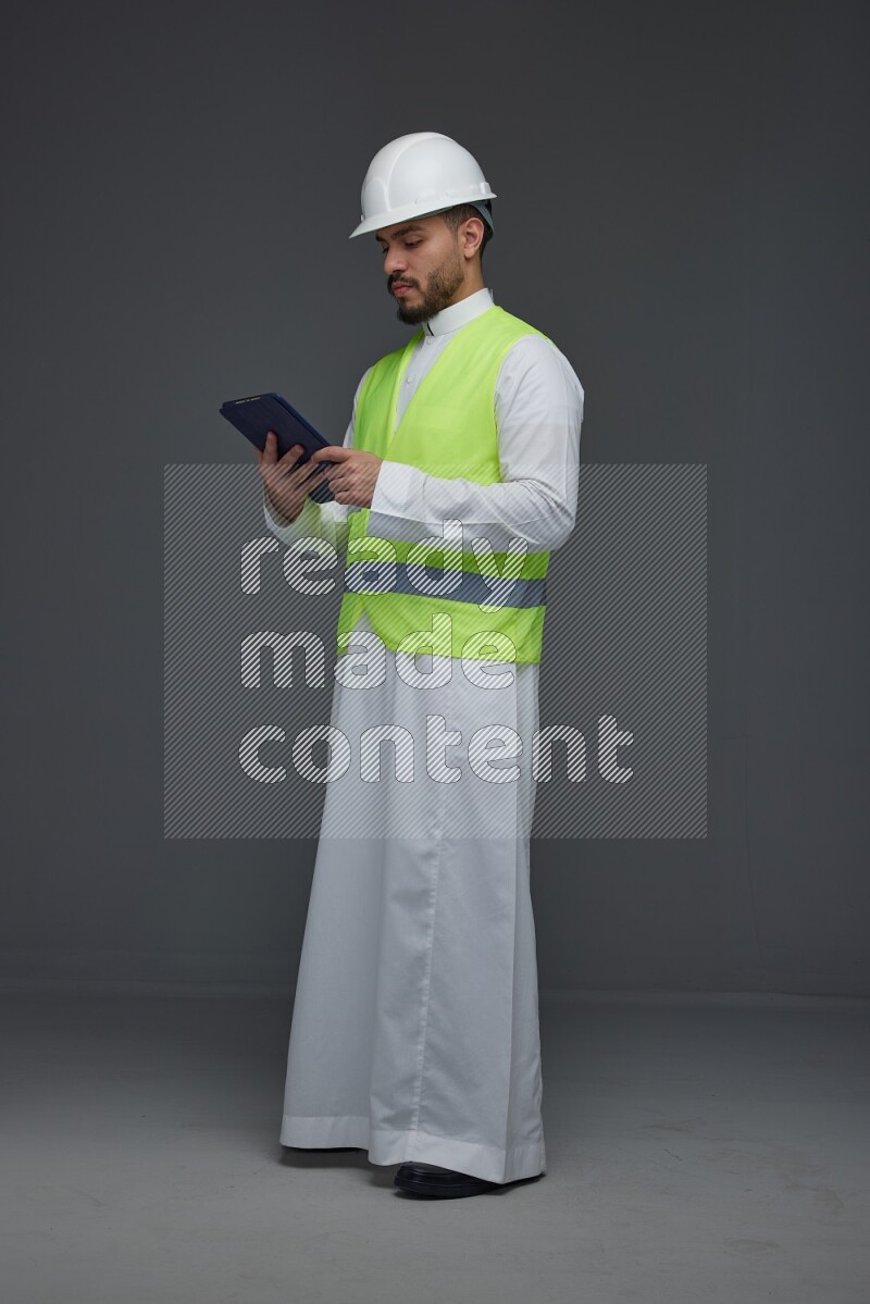 A Saudi man wearing Thobe with a yellow safety vest and white helmet standing and using his tablet different angles eye level on a gray background