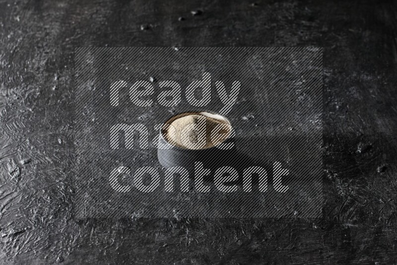 Black pottery bowl full of white pepper powder on textured black flooring