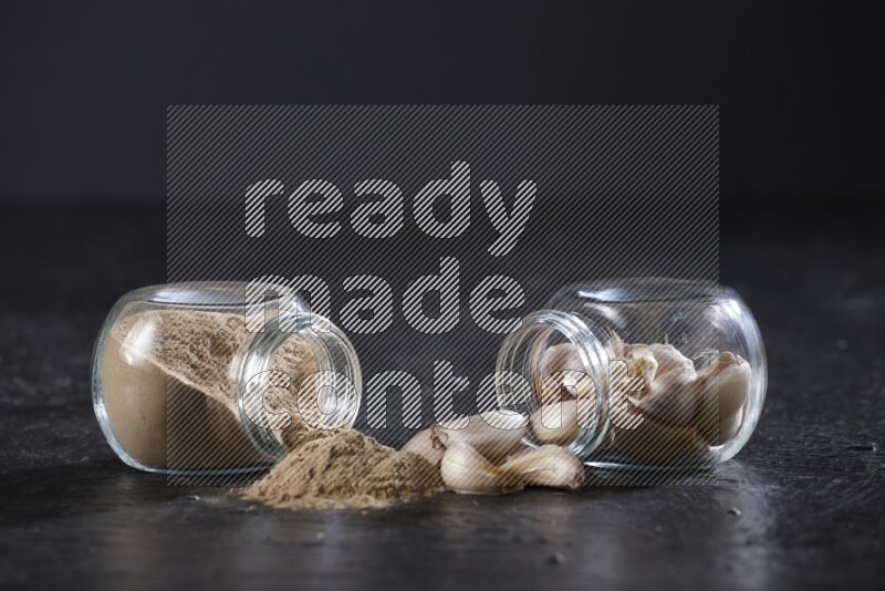 2 glass spice jars, one full of garlic powder and the other full of garlic cloves both flipped on a textured black flooring