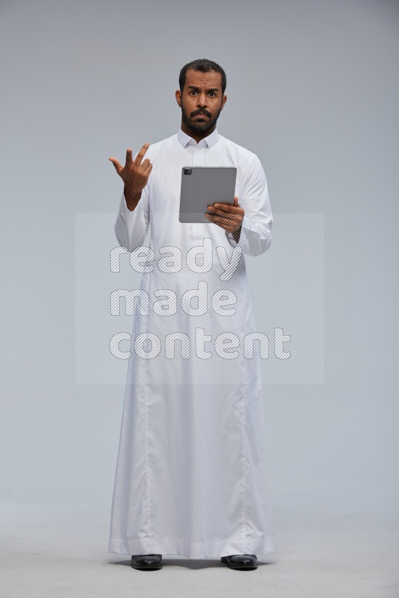 Saudi man wearing Thob standing working on tablet on Gray background