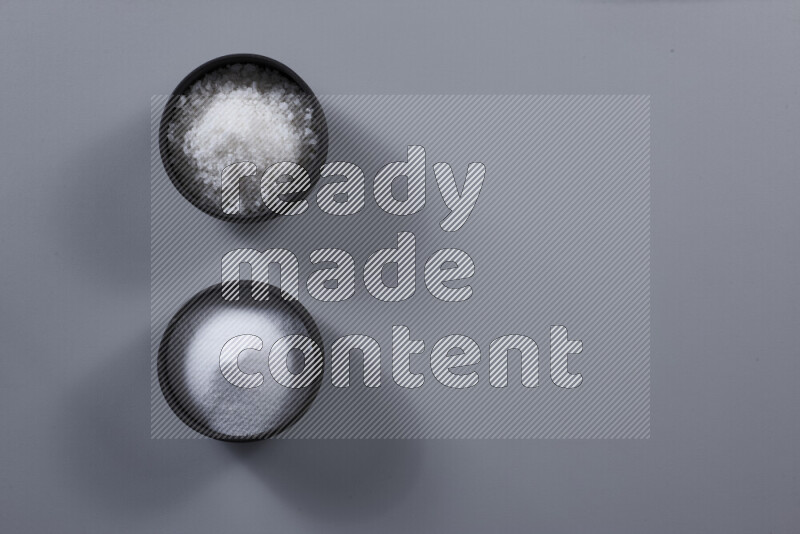 2 pottery bowls one is filled with table salt and the other with coarse sea salt on grey background