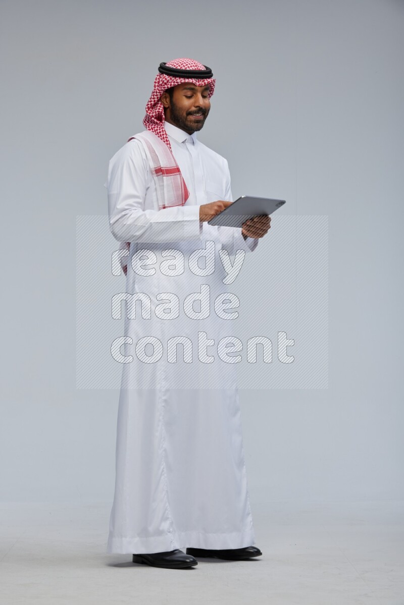 Saudi man Wearing Thob and shomag standing working on tablet on Gray background