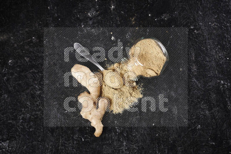 A glass jar full of ground ginger powder flipped with some spilling powder on black background