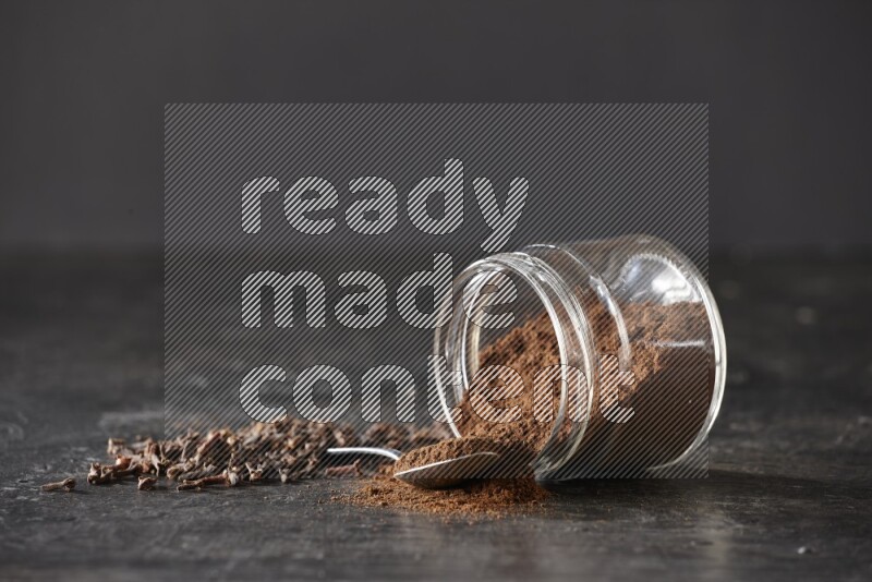 A flipped glass jar and metal spoon full of cloves powder with cloves spread on a textured black flooring