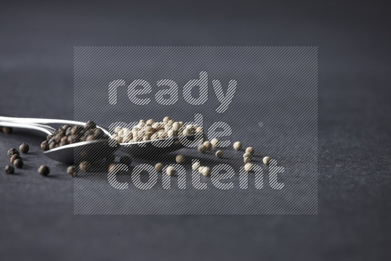 ملعقتان معدنيتان ممتلئتان بحبوب الفلفل الأسود والأبيض مع حبوب متناثرة على أرضية سوداء