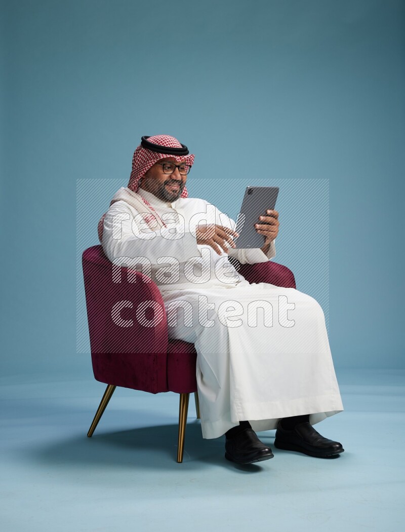 Saudi Man with shimag sitting on chair working on tablet on blue background