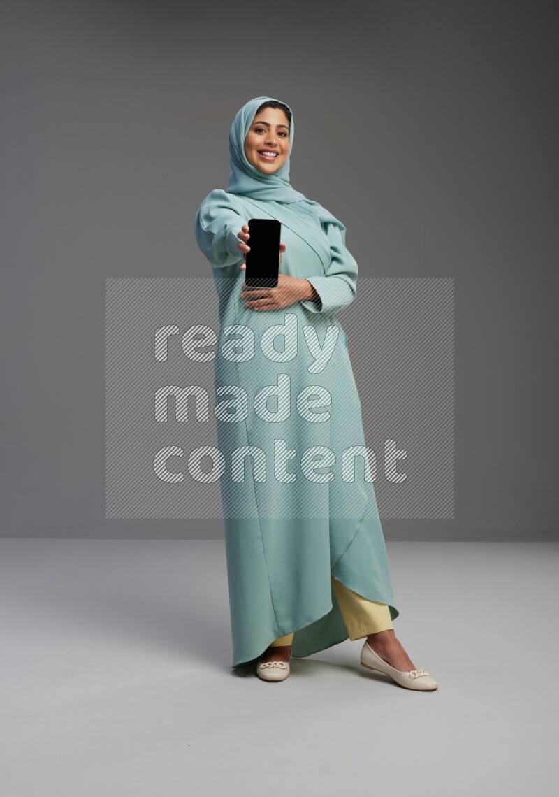 Saudi Woman wearing Abaya standing showing phone to camera on Gray background