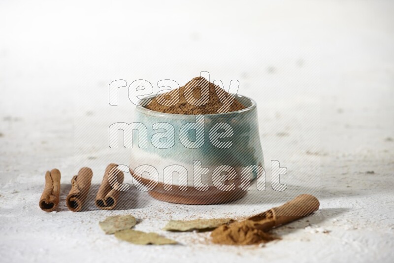 Cinnamon powder in a ceramic bowl with cinnamon sticks and laurel leaves on white background