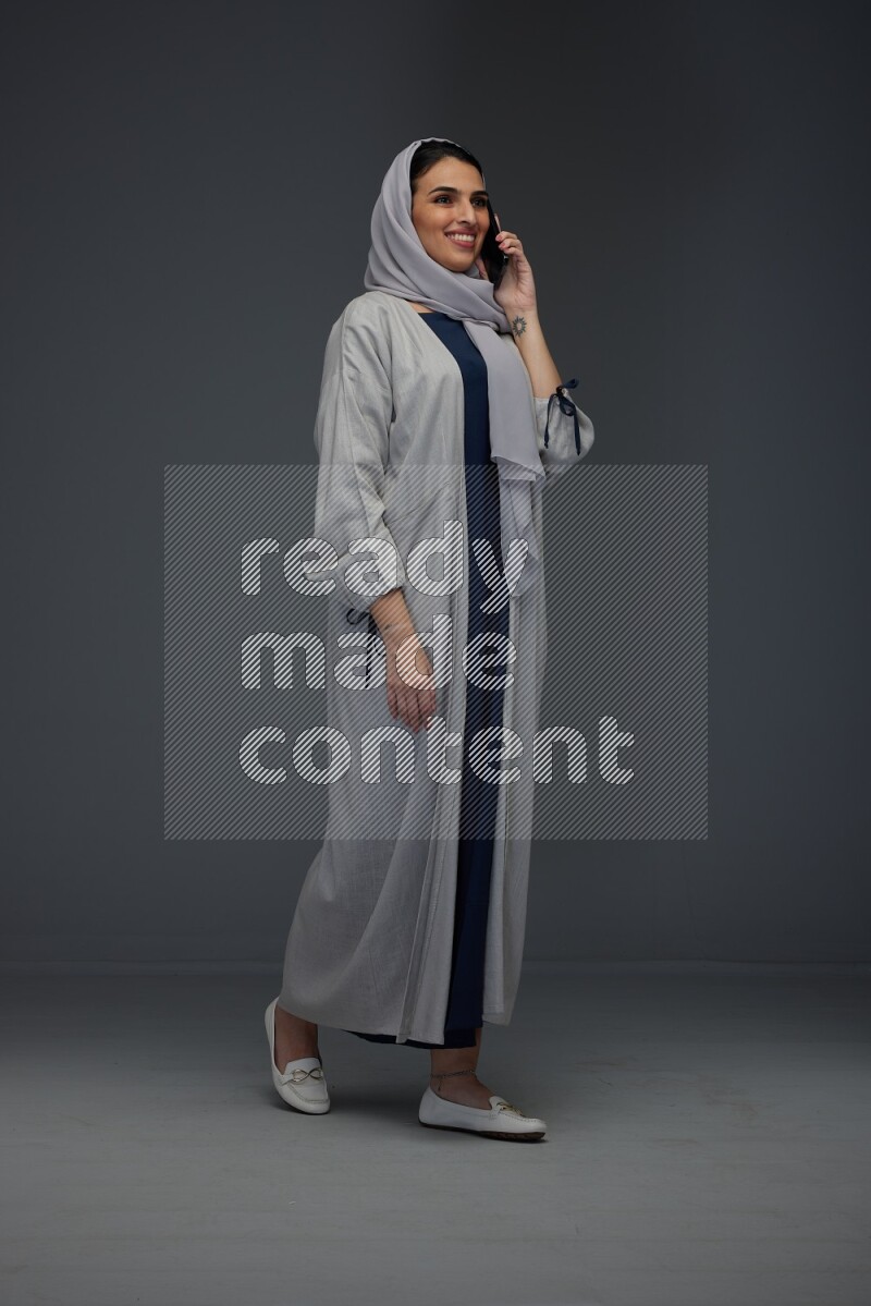 A Saudi woman wearing a light gray Abaya and head scarf standing and talking in the phone eye level on a grey background