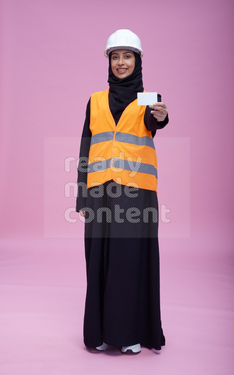 Saudi woman wearing Abaya with engineer vest and helmet standing holding ATM card on pink background