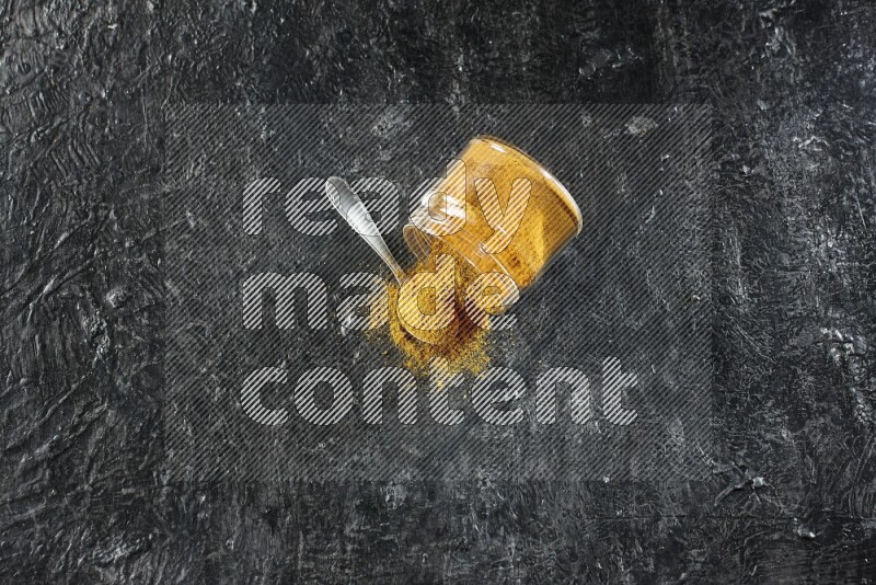 A flipped glass jar and a metal spoon full of turmeric powder and powder spilled out of it on textured black flooring