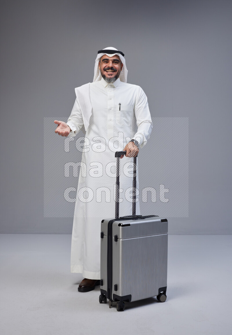 Saudi man wearing Thob and white Shomag standing holding Travel bag on Gray background