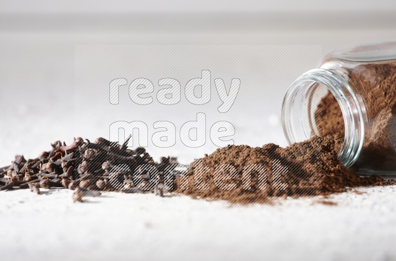 A glass spice jar full of cloves powder flipped and powder came out of it with cloves spread on textured white flooring