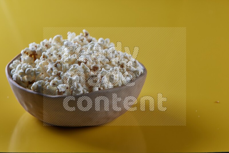 A brown pottery bowl full of popcorn on a yellow background in different angles