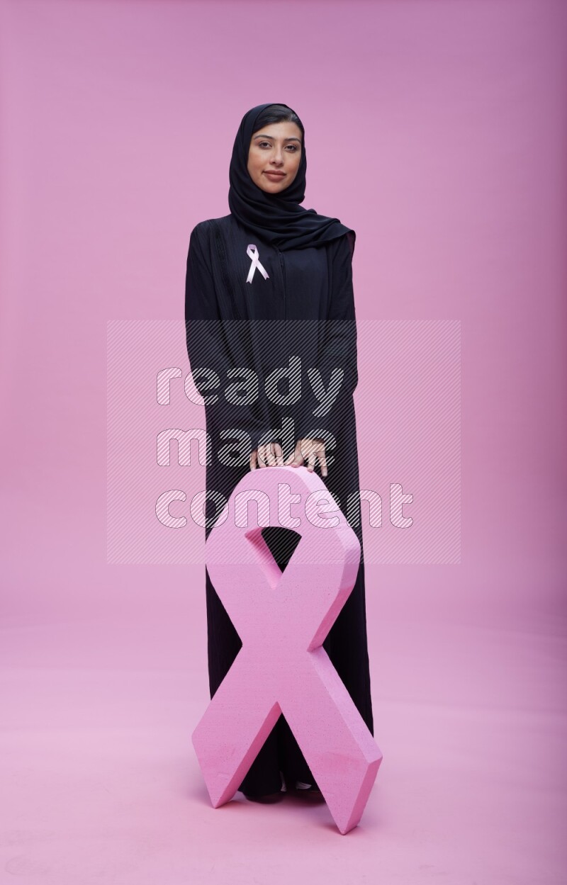 Saudi woman wearing pink ribbon on abaya standing holding awareness ribbon on plain pink background