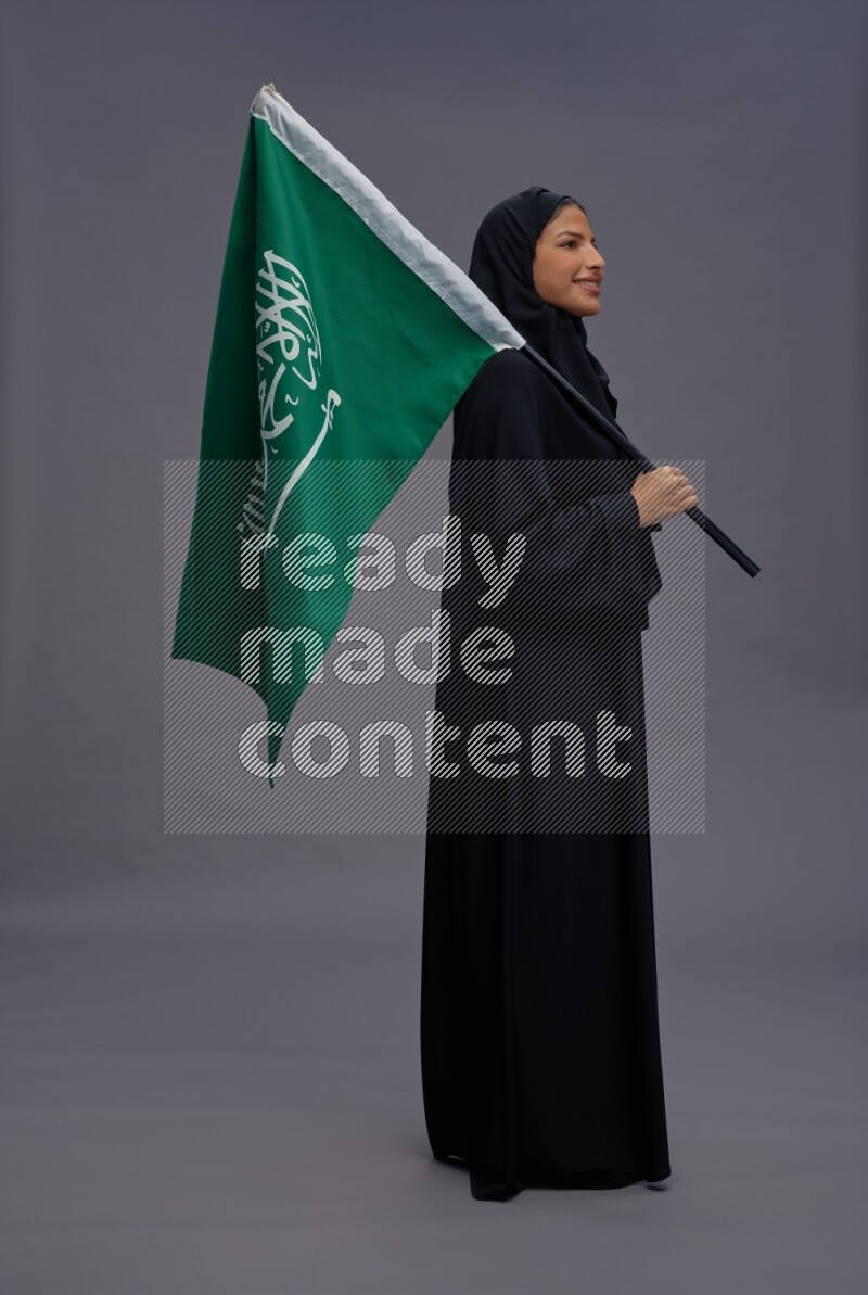 Saudi woman wearing Abaya standing holding Saudi flag on gray background