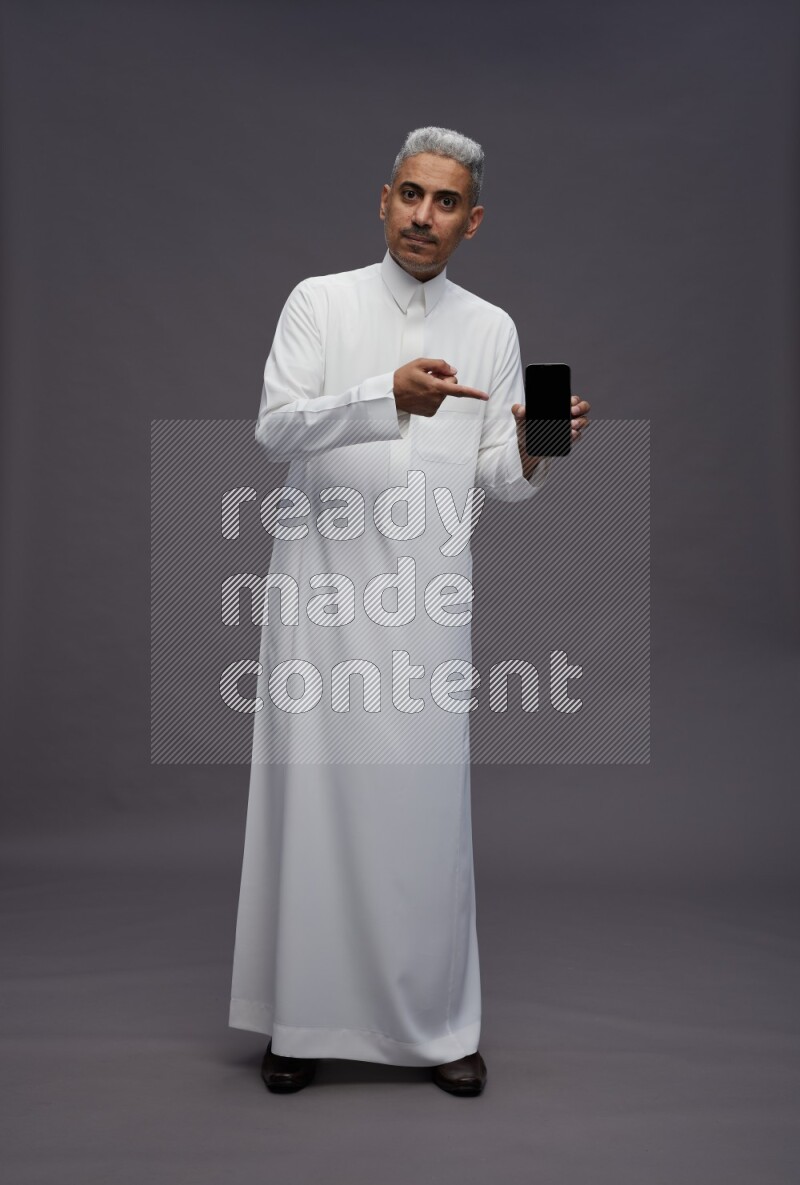 Saudi man wearing thob standing showing phone to camera on gray background