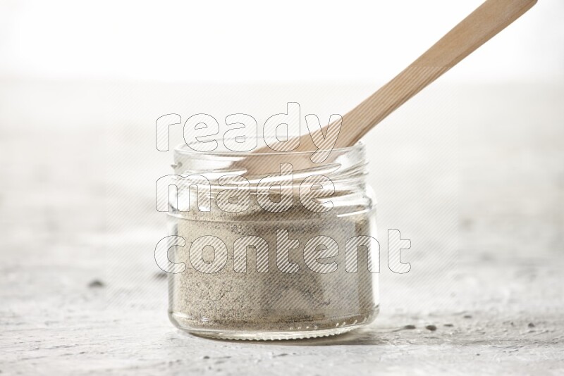 A glass jar and wooden spoon full of white pepper powder on textured white flooring
