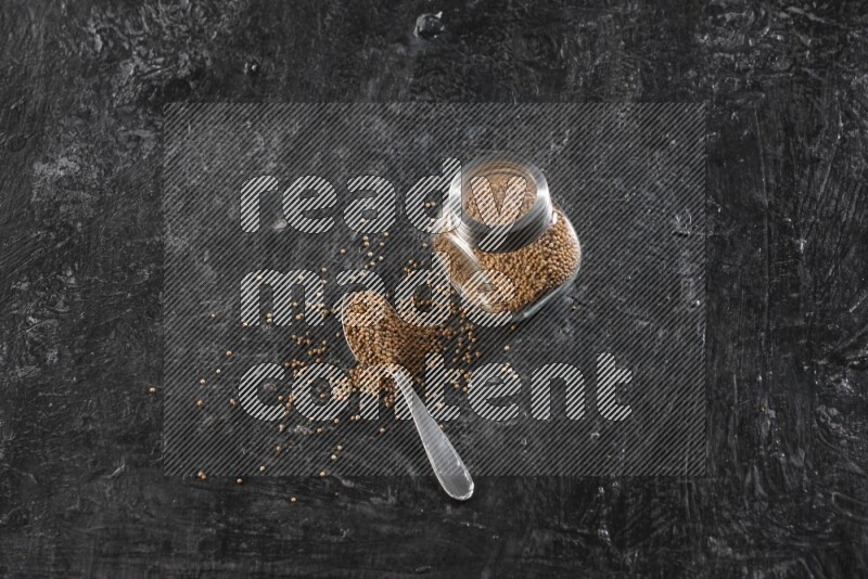 وعاء زجاجي وملعقة معدنية كلاهما ممتلئ ببذور الخردل على أرضية سوداء