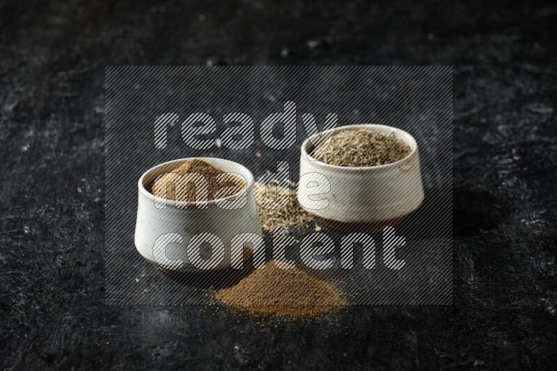2 beige bowls, one full of cumin powder and the other full of seeds on a textured black flooring
