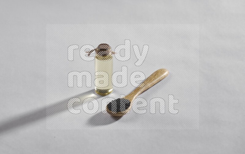 A wooden spoon full of black seeds with a glass bottle of black seeds oil on a white flooring