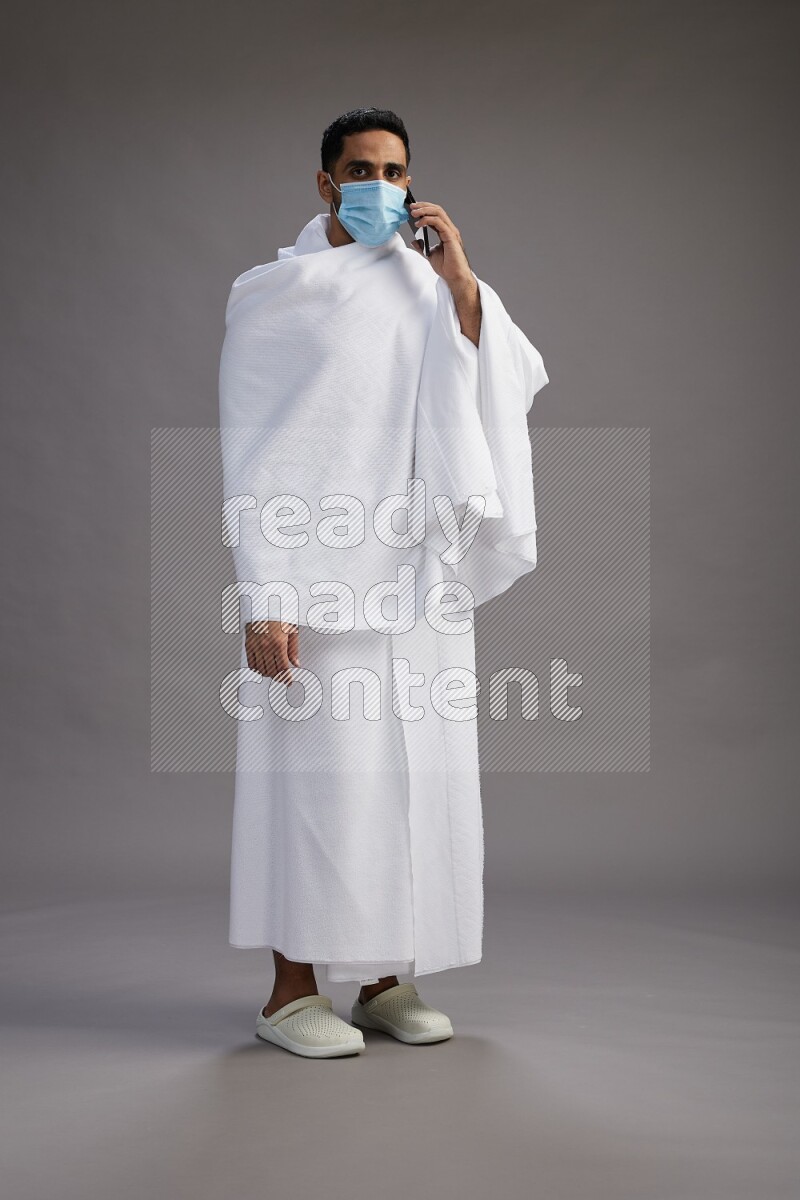 A man wearing Ehram with face mask Standing talking on phone on gray background