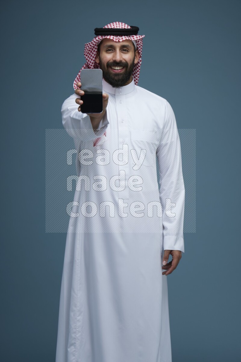 A man showing his phone Screen in a blue background wearing Saudi Thob with Shomag