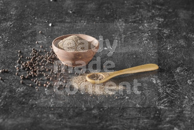 A wooden bowl and a wooden spoon full of black pepper powder with the beads on a textured black flooring