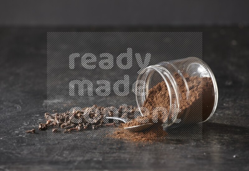 A flipped glass jar and metal spoon full of cloves powder with cloves spread on a textured black flooring