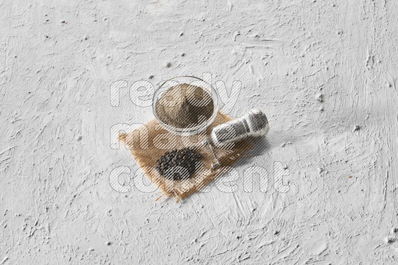 A glass bowl full of black pepper powder, black pepper beads and a turkish metal grinder on burlap fabric on textured white flooring